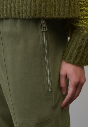 Primer plano de una mano descansando al lado de una pierna de pantalón verde con un zipper plateado vertical y la manga de un suéter verde de punto.