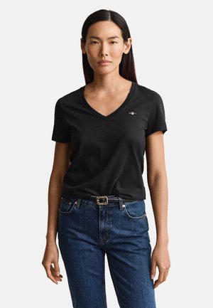 Donna con capelli lunghi e scuri che indossa una maglietta nera con scollo a V e jeans blu con cintura nera, in piedi davanti a uno sfondo semplice.