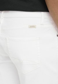 Bijele traperice imaju stražnji džep s bež etiketom na kojoj piše "DEDICATED TO DENIM PERFECTION". Diskretni žuti šavovi naglašavaju.