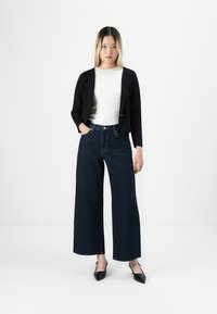 Une jeune femme se tient debout, une main dans la poche, portant une veste noire courte, un haut blanc, un jean large foncé et des talons aiguilles noirs.