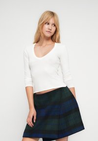 Λευκή μακρυμάνικη μπλούζα με V-λαιμόκοψη, συνδυασμένη με πράσινη και μπλε καρό φούστα σε γραμμή Α. Το ύφασμα φαίνεται απαλό και υφασμάτινο.