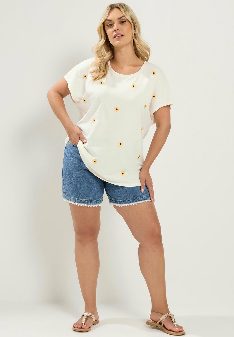 Woman with long blonde hair wearing a white t-shirt with small yellow flowers, blue denim shorts with lace trim, and beige sandals.
