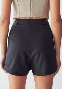 Černé šortky vyrobené z hladkého, lehkého materiálu. Mají čistý, přesně střižený design s vysokým pasem a postranními kapsami.