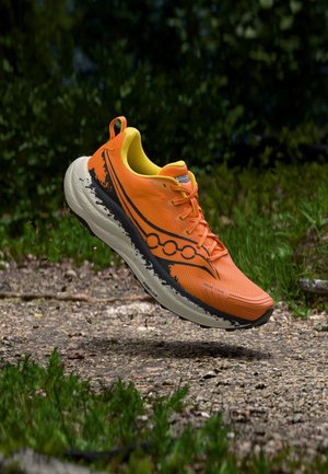 Leuchtend orangefarbener Trailrunning-Schuh mit schwarzer und weißer Sohle in der Luft über einem Kiesweg mit grünem Laub im Hintergrund.
