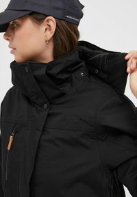 Schwarze wasserdichte Jacke mit hohem Kragen, Druckknopfverschlüssen, Reißverschluss-Tasche und Logodetail. Glatte Textur mit minimalen Designelementen.
