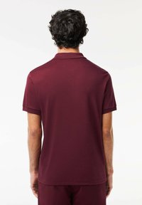 Burgundska polo majica s kratkimi rokavi, standardnim ovratnikom in gladko teksturo, videna s hrbtne strani. Brez vidnih logotipov ali vzorcev.