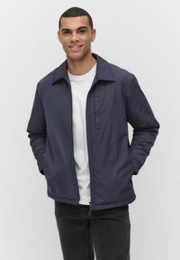 Marinblå tygjacka med krage, dragkedja fram och sidofickor, bärs över en vit t-shirt och kombineras med svarta byxor.