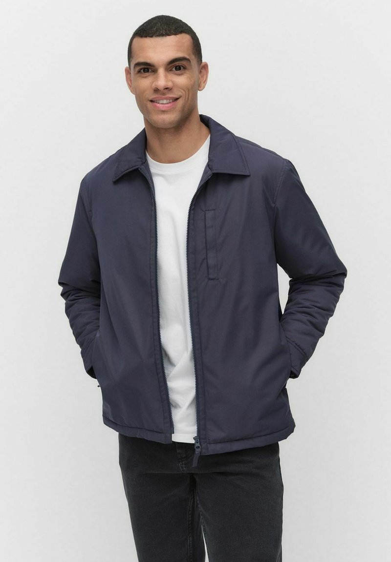 Marinblå tygjacka med krage, dragkedja fram och sidofickor, bärs över en vit t-shirt och kombineras med svarta byxor.