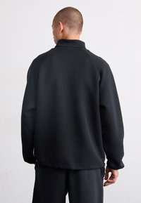 Veste à capuche noire avec un col haut, des manches raglan et une texture douce. Comprend un cordon de serrage au niveau de l'ourlet.