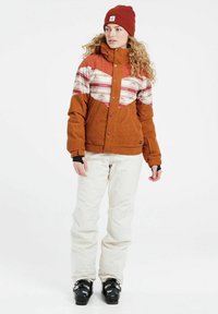 Bruine corduroy jas met een multi-kleurige gestreepte patroon op de borst, gecombineerd met beige broeken en zwarte skibotten. Rode beanie op.