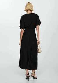 Donna con capelli biondi corti, vestita con un abito midi plissettato nero e sandali neri con tacco alto, mentre tiene in mano una piccola borsa beige con borchie.