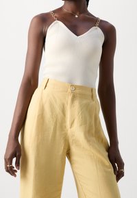 Cream ribbed tank top with chain straps, paired with high-waisted, light yellow wide-leg trousers. Smooth texture, minimalist design.