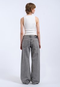 Jean gris à jambe large avec une coupe relax, en denim, et un débardeur ajusté blanc, vu de derrière sur un fond uni.