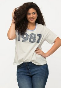 Bílý tričko s krátkým rukávem s velkými vyšitými čísly "1987" z denimu, kombinované s tmavě modrými přiléhavými džínami.