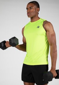 Limoengroene mouwloze sportshirt met een aansluitend ontwerp, gecombineerd met zwarte shorts. Het onderwerp houdt hexagonale dumbbells vast.