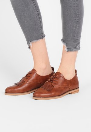 Bruine leren veterschoenen met franjes, gedragen met grijze cropped jeans op een lichte achtergrond.