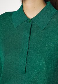 Gros plan d'une personne portant un haut en maille vert à col avec boutons, à texture légèrement brillante.