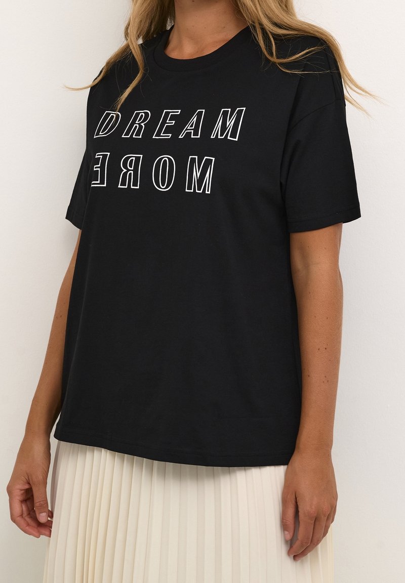 Femme portant un t-shirt noir avec le texte "DREAM MORE" en contour blanc et une jupe plissée blanche sur un fond blanc uni.
