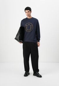 Sudadera azul marino con un diseño de cara sonriente y texto, combinada con pantalones negros holgados y zapatos negros, sosteniendo un bolso negro.