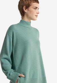 Kobieta z krótkimi włosami, ubrana w jasnoniebieski sweter z golfem z długimi rękawami i prążkowanymi krawędziami, patrzy w bok, trzymając rękę w kieszeni.