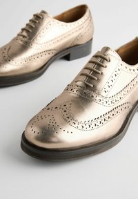 Chaussures oxford en bronze métallique avec des détails en pointe de aile, des perforations, et une semelle en caoutchouc noir légèrement surélevée. Les lacets courent le long du dessus.