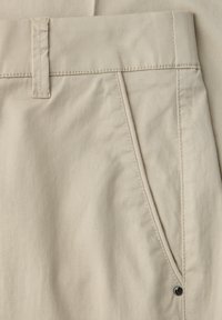 Zbliżenie na beżowe spodnie pokazujące szlufkę na pasek, przednią kieszeń z metalowym ćwiekem oraz przeszycia na bawełnianym materiale.