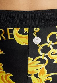 Crne tajice s uzorkom i žutim baroknim dizajnom, s teksturiranim pojasom na kojem piše "VERSACE" i ukrasnim motivom lanca.
