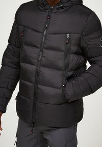 Schwarze Steppjacke mit Kapuze, gestepptem Design, zwei Brusttaschen mit Reißverschluss und Seitentaschen. Mit kontrastierenden Reißverschlüssen und verstellbarem Kordelzug.