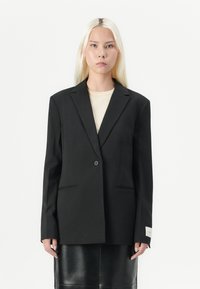 Czarna, oversize marynarka z wyraźnymi ramionami, zapinana na jeden guzik i z bocznymi kieszeniami. Wykonana z gładkiego materiału, z kremową metką na rękawie.