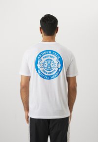 Balts īsām piedurknēm T-krekls ar lielu zilu grafiku mugurpusē, kurā ir uzraksts "ARMOUR CLUB" un zeme, ko ieskauj izlocītas rokas.