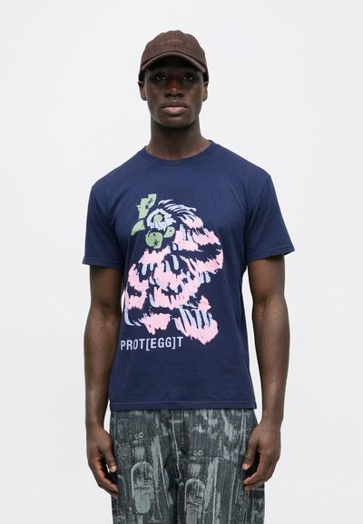 Μπλε ναυτικό βαμβακερό μπλουζάκι με πολύχρωμο γραφικό τύπωμα χαρακτήρα και το κείμενο «PROT[EGG]T», συνδυασμένο με γκρι παντελόνι με υφασμένο σχέδιο.