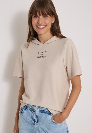 Femme souriante aux cheveux bruns portant un sweat à capuche beige à manches courtes avec le texte "SUN KNOWS THE WAY" et un jean bleu clair, main dans la poche.