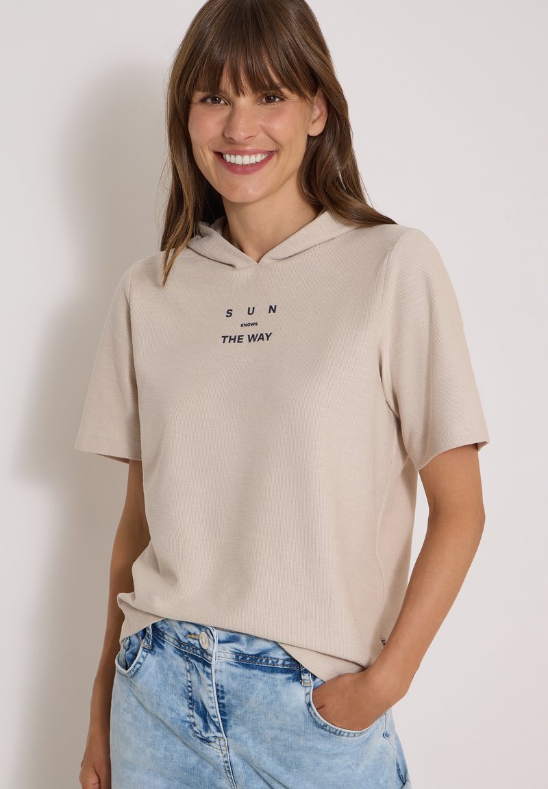 Femme souriante aux cheveux bruns portant un sweat à capuche beige à manches courtes avec le texte "SUN KNOWS THE WAY" et un jean bleu clair, main dans la poche.