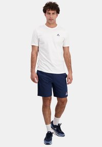 T-shirt bianco a maniche corte con logo, abbinato a pantaloni corti blu navy. Il modello indossa scarpe da ginnastica blu navy e grigie con calze bianche.