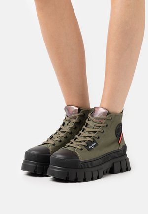 Baskets montantes vertes en toile avec un bout en caoutchouc noir, une semelle chunky noire et des œillets en métal ; elles comportent un patch logo et des lacets.