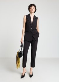 Blazer noir sans manches avec un profond décolleté en V et une fermeture à un bouton, associé à un pantalon noir fuselé ; accessoires comprenant un sac noir et une écharpe jaune.