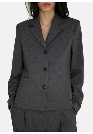 Kobieta ubrana w ciemnoszary, dopasowany żakiet z trzema guzikami i pasujące spodnie, ręce w kieszeniach, na jednolitym tle.