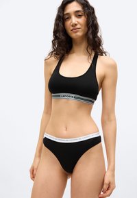 Woman standing with hands relaxed, wearing black sports bra and matching black briefs with white branded waistbands against plain background.