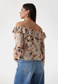 Floral off-the-shoulder top with ruffle detailing, featuring warm hues of beige, brown, and teal, paired with light blue high-waisted jeans.