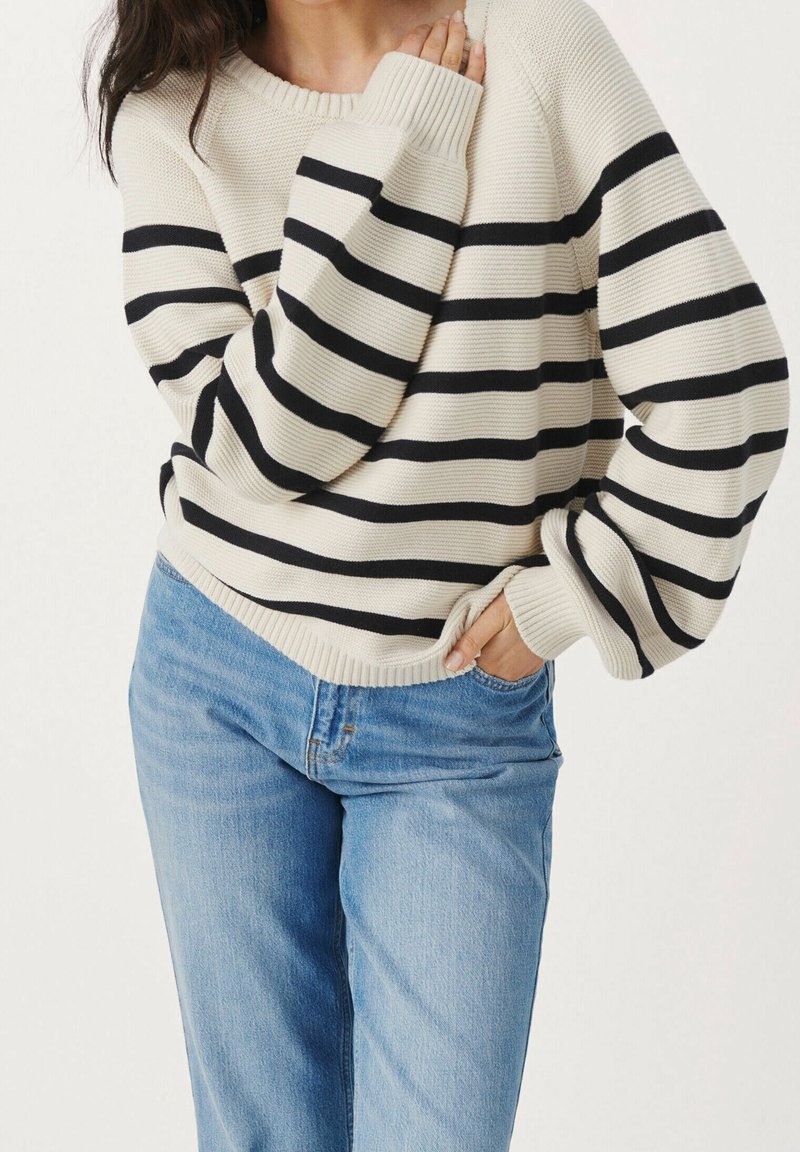 Personne portant un pull beige avec des rayures horizontales noires et un jean bleu clair, une main dans la poche, l’autre près de la clavicule.
