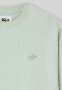 Sweatshirt surdimensionné vert clair avec un col rond, arborant un subtil design de sneaker brodé à l'avant. Tissu doux et texturé.
