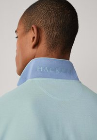 Polo tričko v světle modré barvě s tmavě modrým límečkem. Límec je zdoben vyraženým nápisem "HACKETT". Hladká textilie, volný střih.