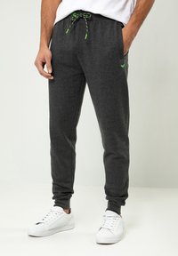Graue Sweatpants mit einem Kordelzugbund, ausgestattet mit grünen Akzenten, seitlichen Taschen und einem schmal zulaufenden Bein, kombiniert mit weißen Sneakern.