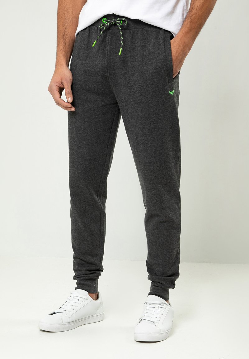 Graue Sweatpants mit einem Kordelzugbund, ausgestattet mit grünen Akzenten, seitlichen Taschen und einem schmal zulaufenden Bein, kombiniert mit weißen Sneakern.