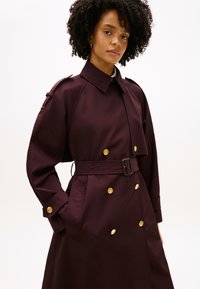 Μπορντό καμπαρντίνα από απαλό ύφασμα, με σχεδίαση διπλής σειράς κουμπιών, χρυσά κουμπιά και ζώνη στη μέση με πλατύ γιακά.