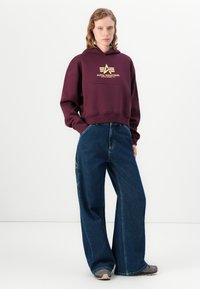 Felpa bordeaux con coulisse e logo dorato sul petto, abbinata a jeans in denim blu a gamba larga. Il tessuto è morbido e ha una vestibilità comoda.