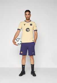 Gul ribbet fodboldtrøje med sorte logoer, lilla shorts, sorte sokker og sorte støvler. Individet holder en mønstret fodbold.