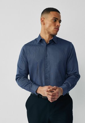 Man met een blauw, lang mouwen, patroonrijke overhemd met knopen, zwarte broek en een zwarte riem, die opzij kijkt met gevouwen handen.