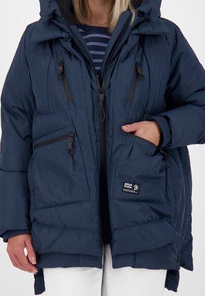 Vrouw draagt een donkerblauw gewatteerd jack met meerdere ritszakken, een capuchon en daaronder een gestreept shirt.