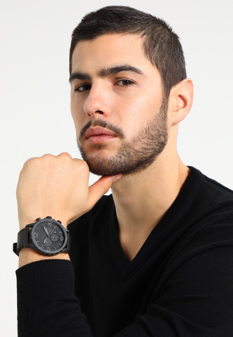 Montre noire avec un cadran rond, marquage blanc contrastant et un bracelet texturé, portée à un poignet, avec une personne reposant son menton sur sa main.